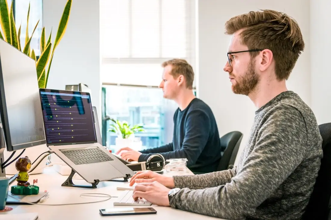 Two developers working at desks with laptops and a large monitor in a bright office